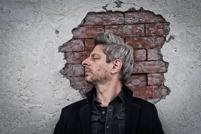 A man in profile before a plaster wall broken open to the brick beneath